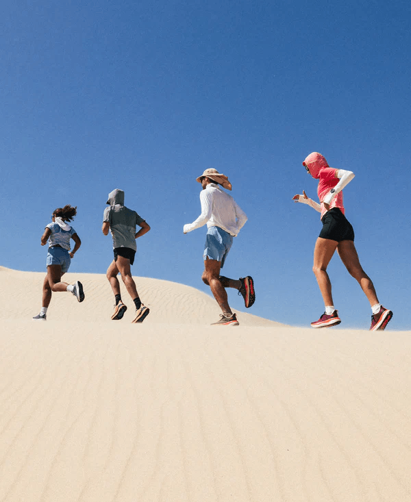 Four runners trail each other on a sandy desert dune under a clear blue sky, wearing athletic clothing in various colors including blue, gray, white, pink, and black.