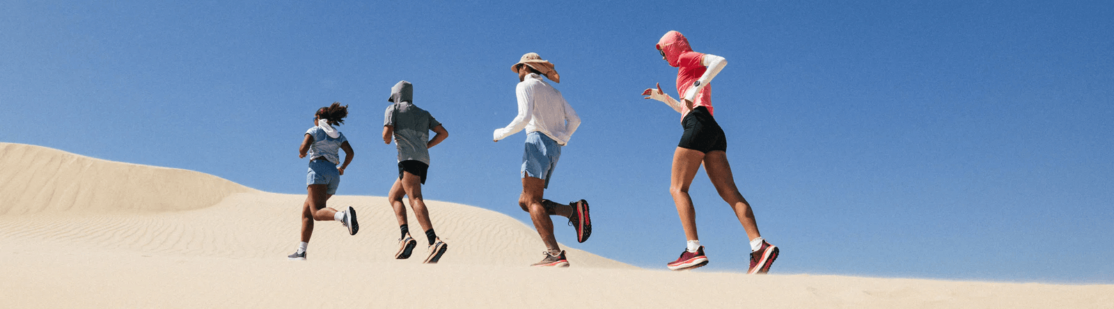 Four people running across sand dunes under a clear blue sky, wearing athletic clothing in various colors including pink, black, white, and gray.