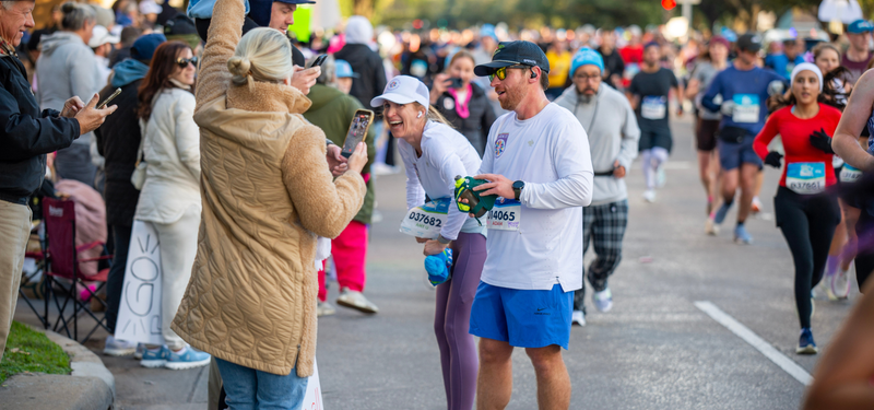 A crowded outdoor marathon scene with runners on a road and spectators lining the street. Two people in the foreground are smiling and taking photos with phones, wearing race bibs and casual athletic clothing.